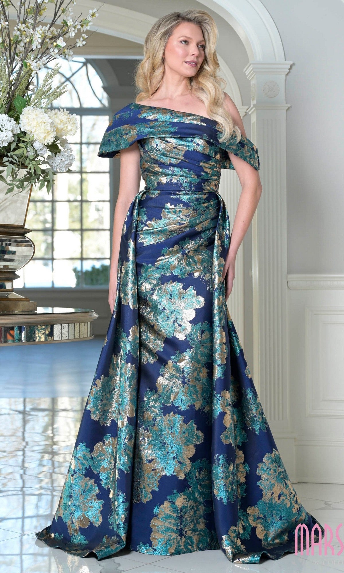 A woman stands indoors near a vase of white flowers, wearing the Marsoni M348 long formal dress in teal multi.